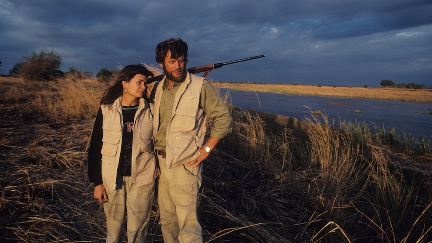 A man with a rifle slung over his shoulders stands on the bank of a river with a woman beside him