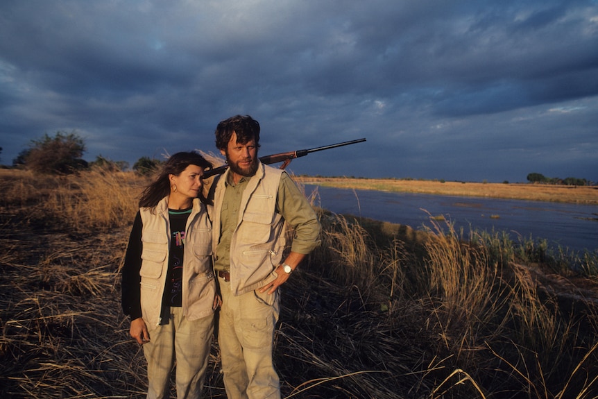 A man with a rifle slung over his shoulders stands on the bank of a river with a woman beside him