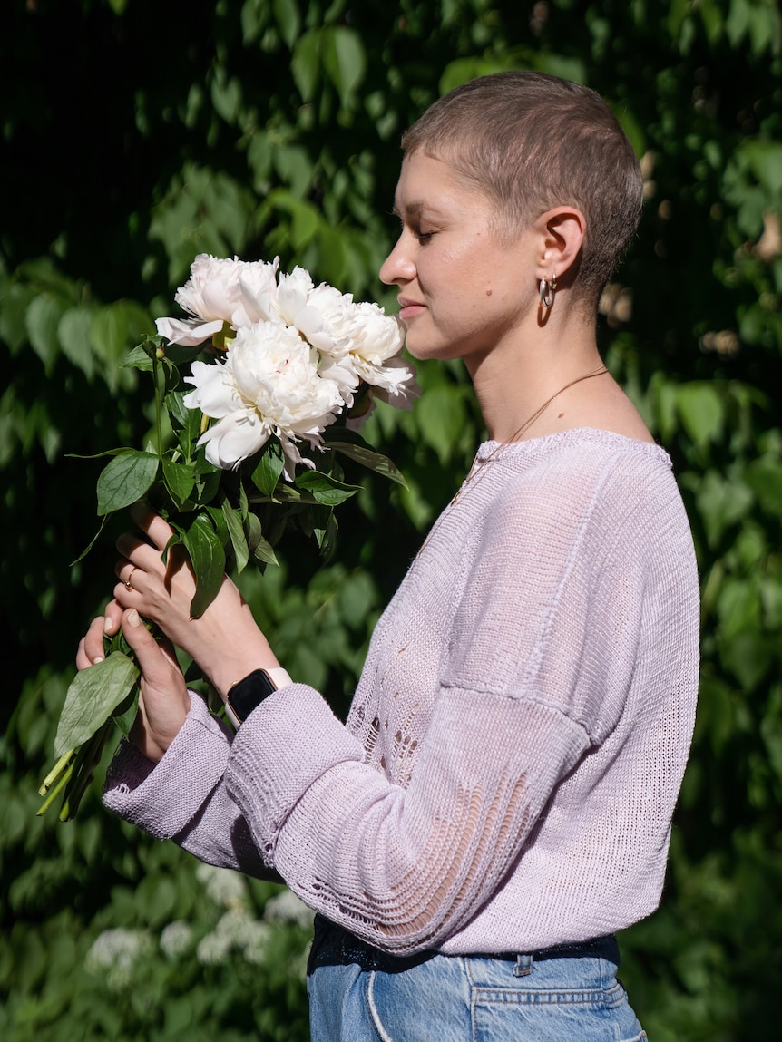 Barbara holds a white bunch of flowers, smelling them with her eyes closed