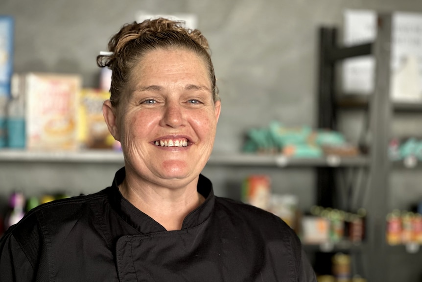 A woman with brown hair and blue eyes smiles, she is wearing a black shirt with shelves of groceries in background