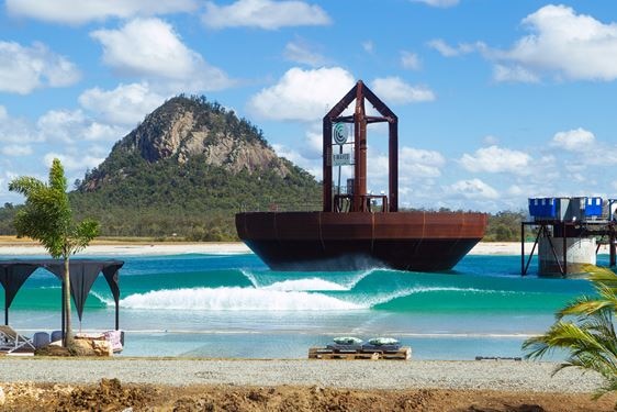 Waves break at a wave pool with palm trees and a mountain surrounding it.