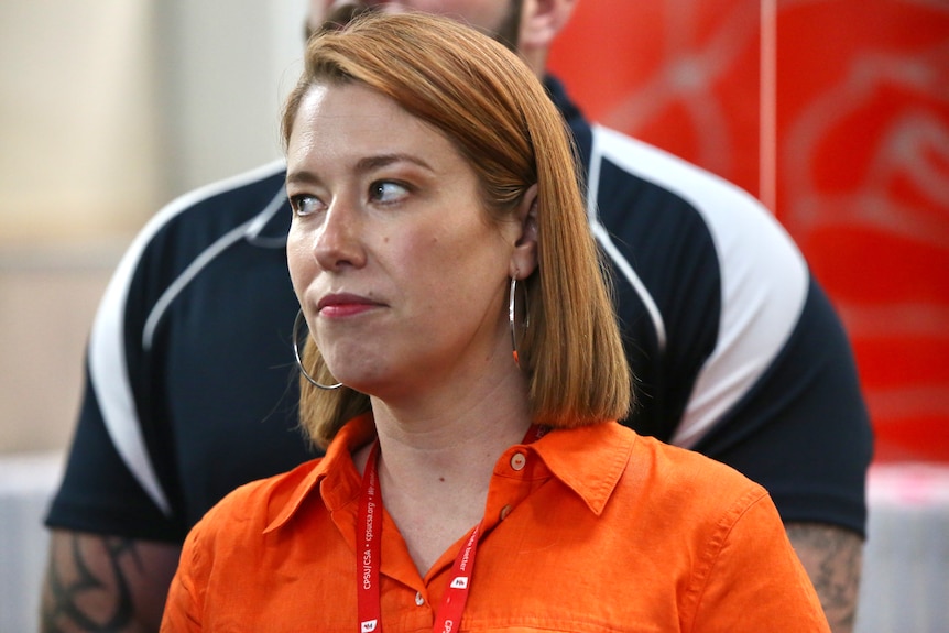 A woman with red hair and an orange shirt looks away off camera.