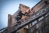 Man in hi-vis and hard hat helps a shirtless man down a ladder 