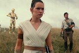Daisy Ridley wears white robes, with other actors standing behind her on a grassy hill.