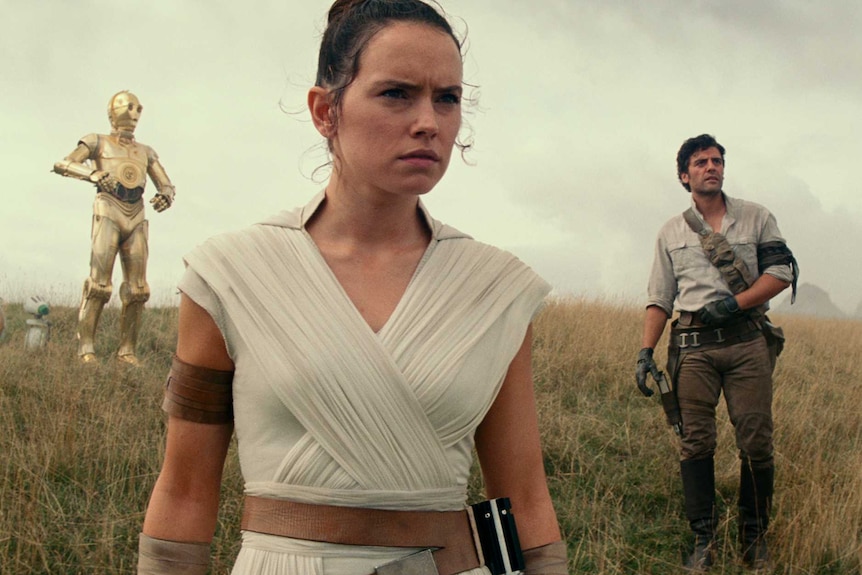 Daisy Ridley wears white robes, with other actors standing behind her on a grassy hill.