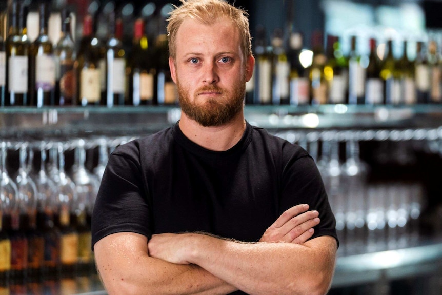 A man in a black shirt stands posing for a photo with his arms folded in front of a bar.