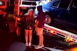 Two young people pose for a photo in front of a car that is loaded onto a tow truck
