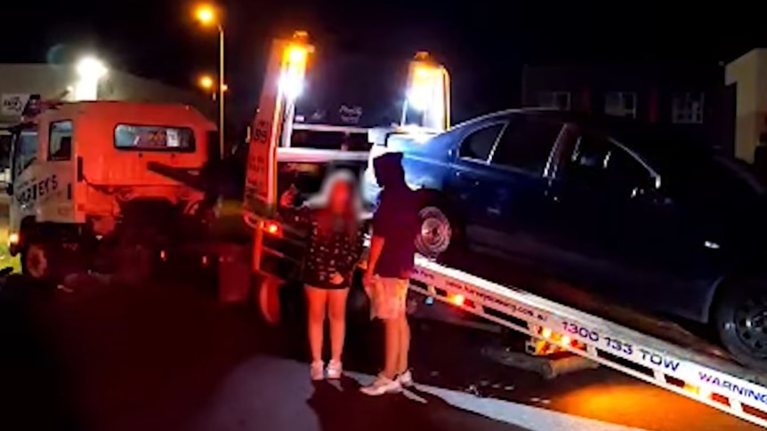 Two young people pose for a photo in front of a car that is loaded onto a tow truck