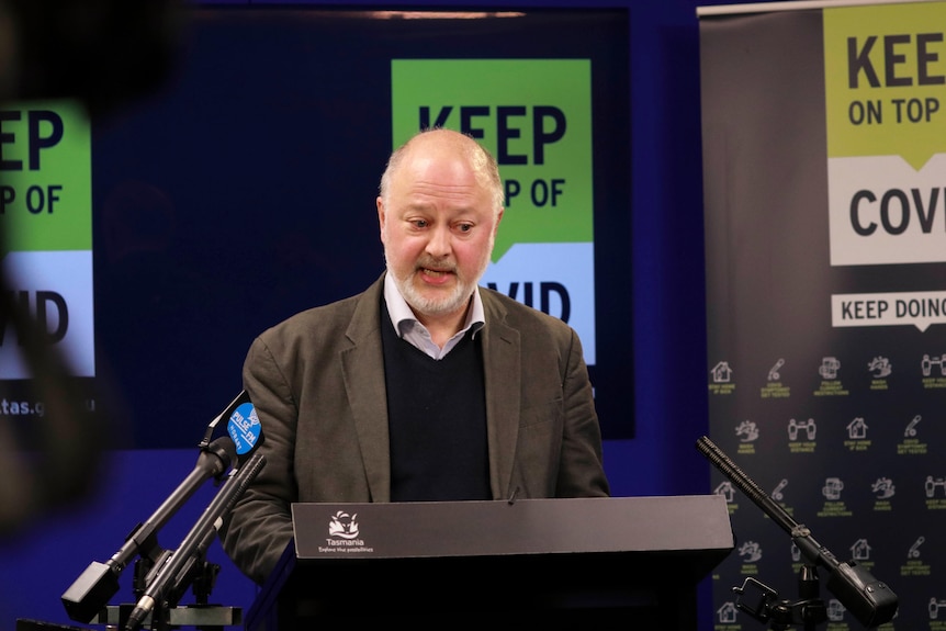 Dr Mark Veitch speaking at a press conference in front of a sign that reads "keep on top of COVID"