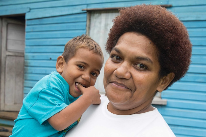 Eseta Liku poses for a portrait holding her small son, as he cheekily puts his thumb in his mouth.