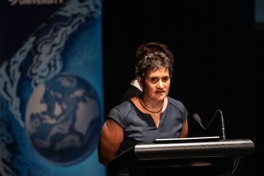 An Indigenous woman stands at a podium in front of a blue background