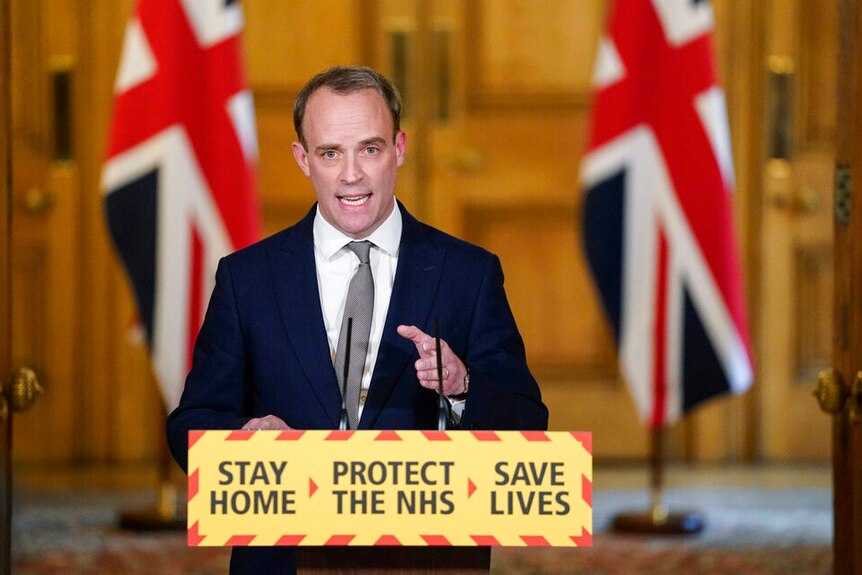 Foreign Secretary Dominic Raab gestures during a coronavirus media briefing standing at a podium in front of two UK flags.