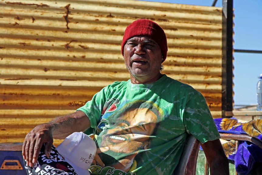Aboriginal man with a green shirt sitting in front of a rusting corrugated iron sheet.