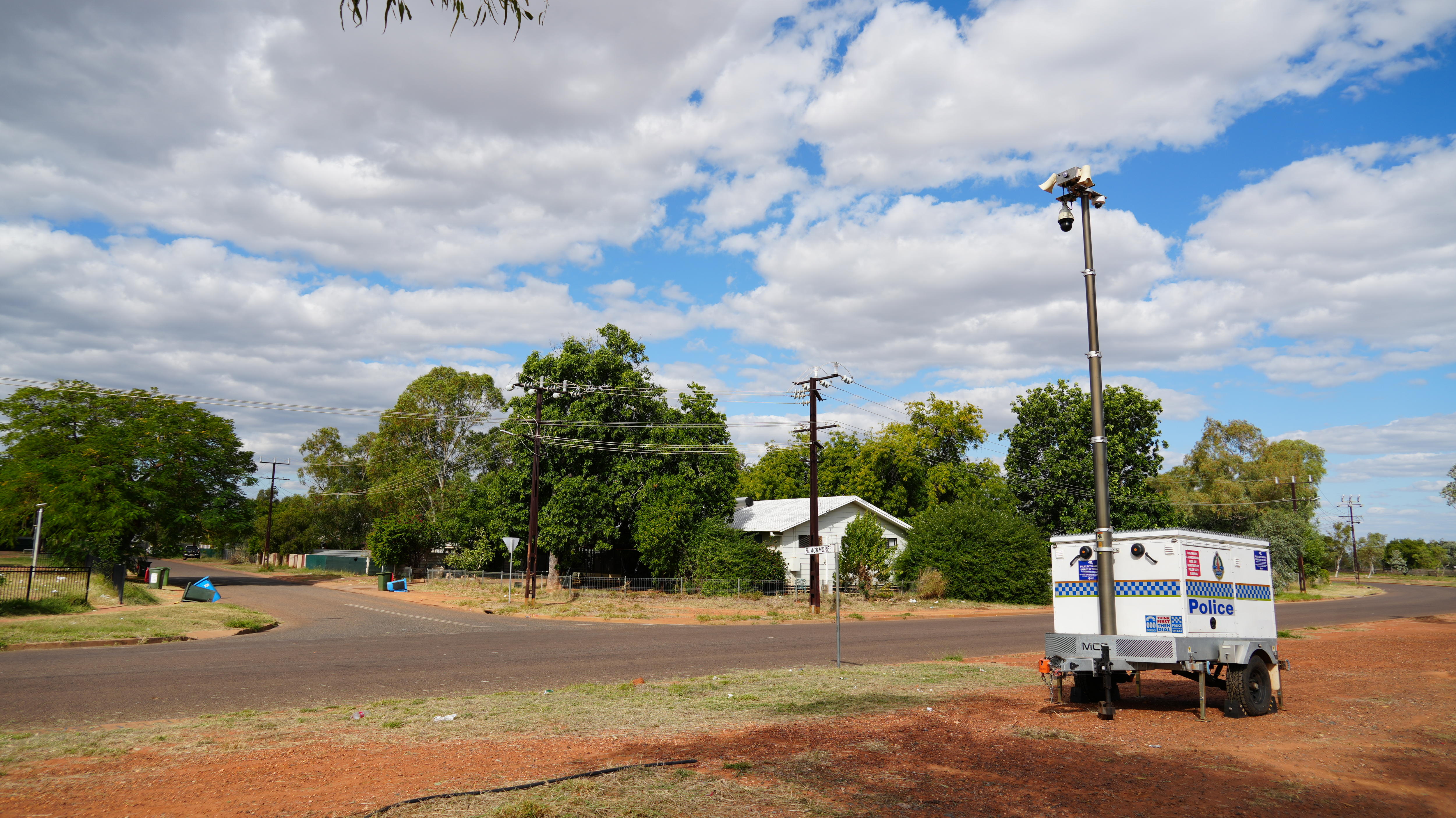 A police storage box unit on the side of the road with security cameras above it, houses in the background.