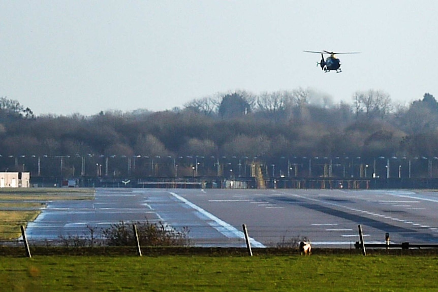 A dark-coloured helicopter hovers over an empty airport runway.