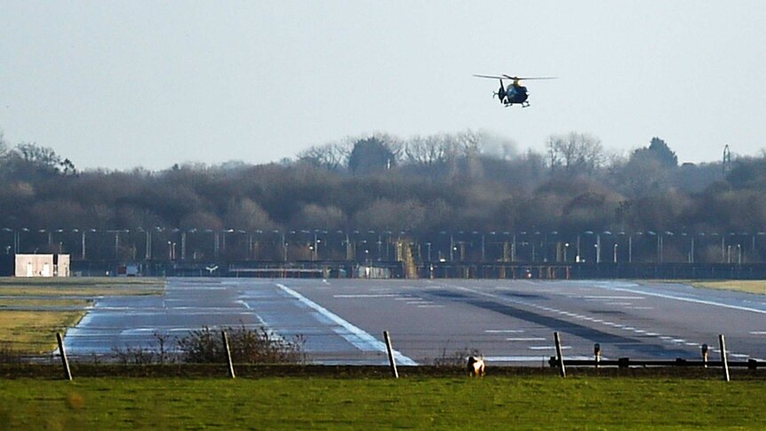 A dark-coloured helicopter hovers over an empty airport runway.