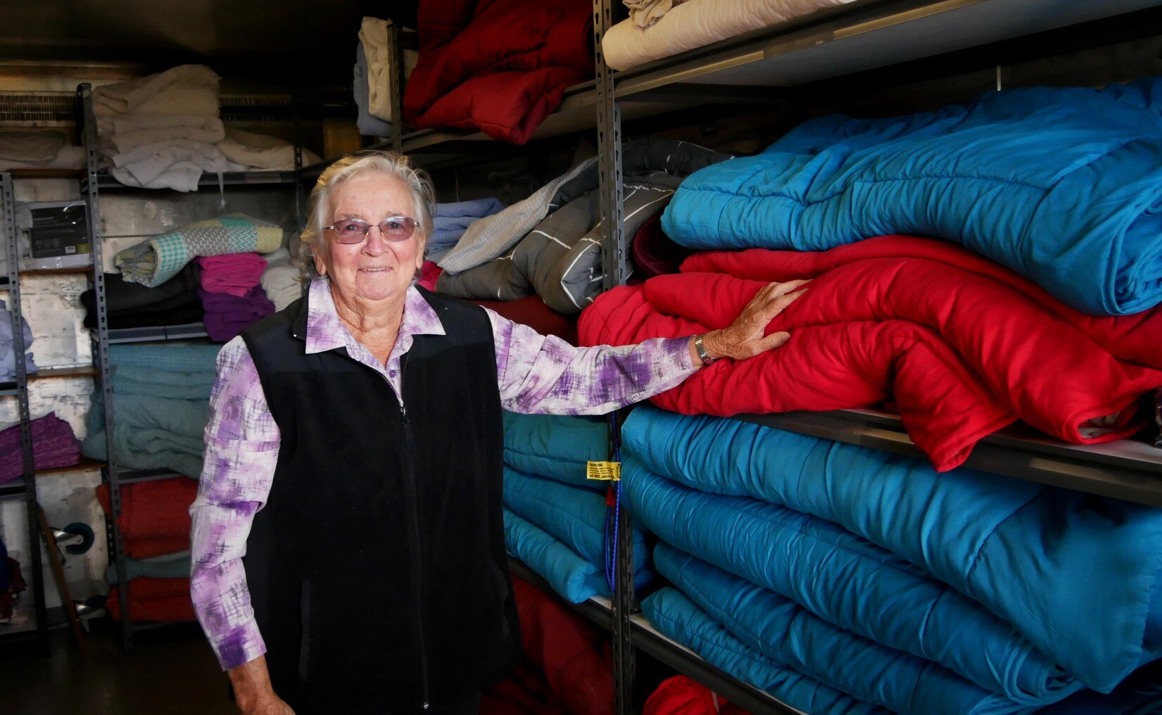An elderly woman with white hair stands in a room lined with shelves full of folded bed linen
