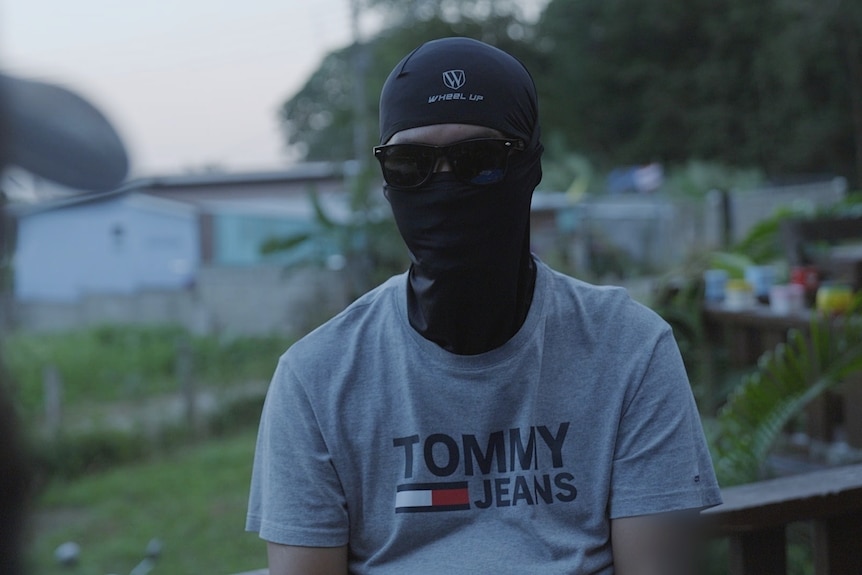 A person in a balaclava and sunglasses with a palmtree in the background.