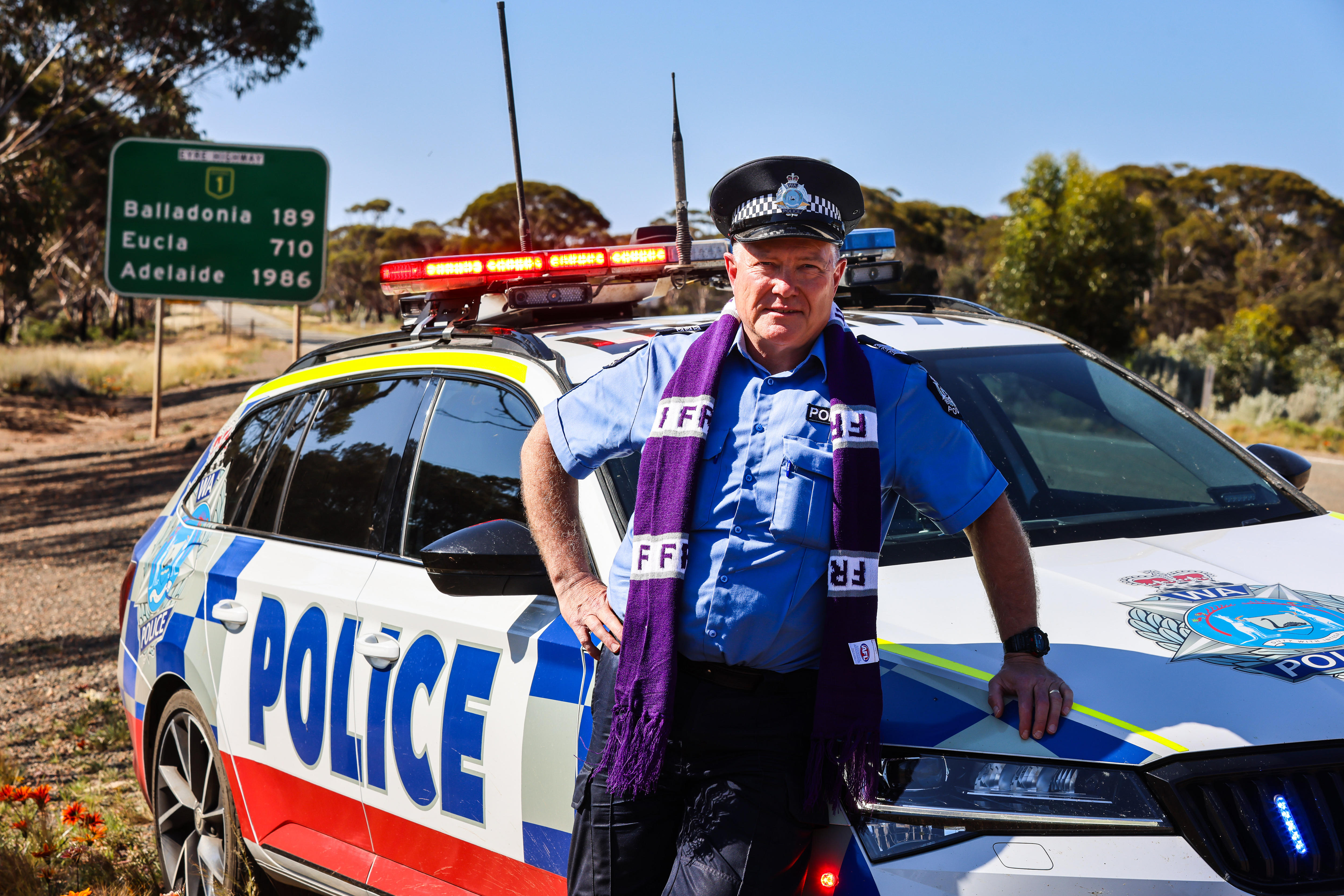 A police officer leaning on a marked car wearing a Fremantle Dockers scarf.
