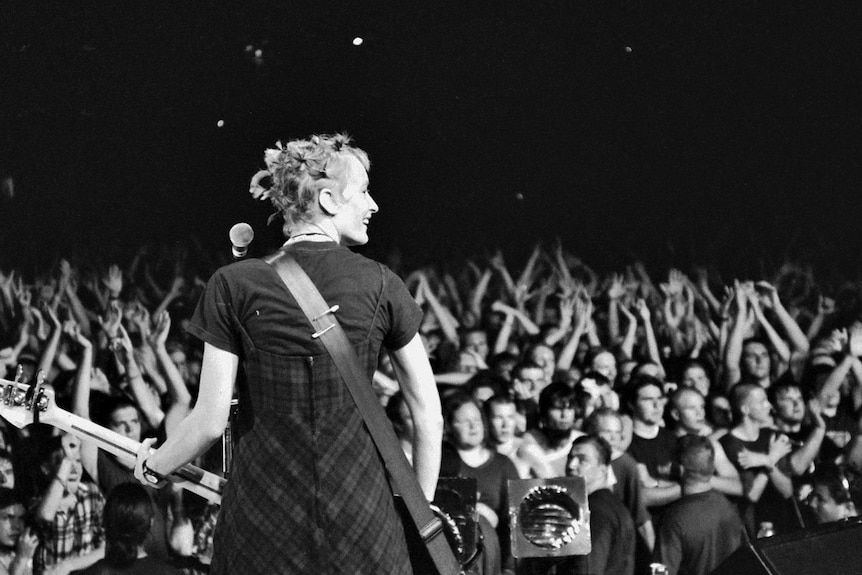 A woman seen from behind smiles holding a guitar on a stage with the crowd in the background