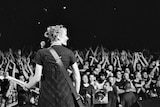 A woman seen from behind smiles holding a guitar on a stage with the crowd in the background