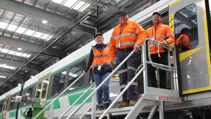 Three workers in high visibility clothing stand on a ramp to the doorway of a new Metronet train.