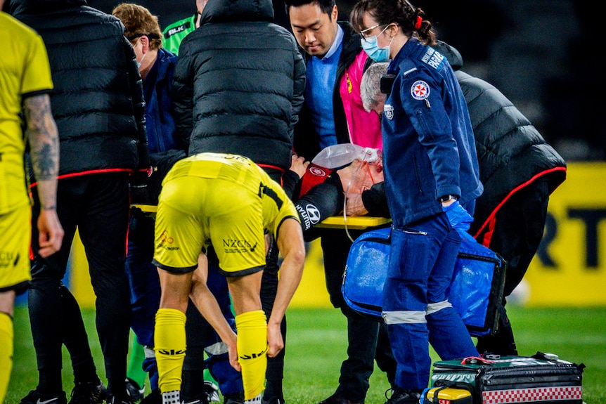 A number of medical workers gather round a footballer on a stretcher as someone checks his neck.