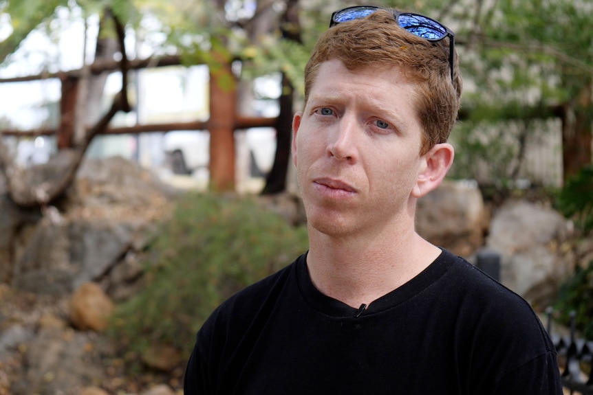 Shoulders and head shot of Mitch Brown looking at the camera, straight face, sunglasses on head, greenery behind.