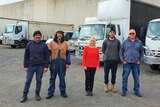 Four men and a woman stand in front of a row of trucks. They are wearing work clothes and boots, expect for a the woman in the m