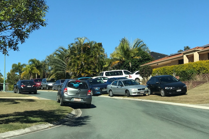 Multiple cars parked on a curb and verge in front of houses on a suburban street.
