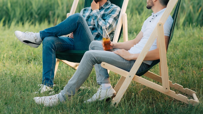 Two men chatting and holding beers while sitting on deck chairs in the grass in front of a field.