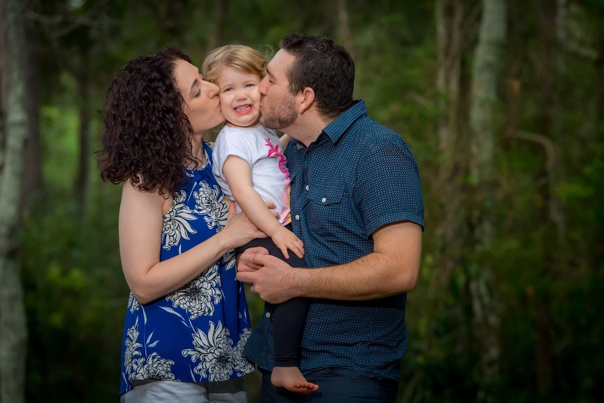 A mother and father hold their young daughter between them, kissing her cheeks.