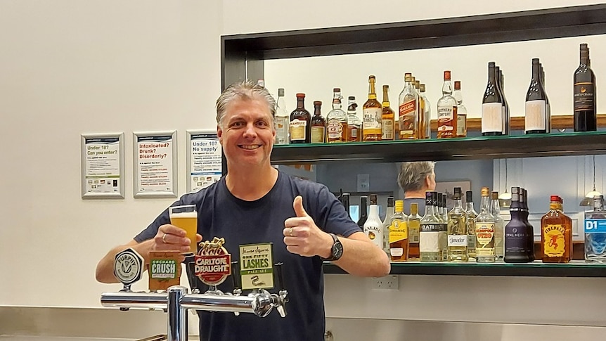 A man stands behind a bar smiling, while holding a beer.