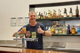 A man stands behind a bar smiling, while holding a beer.