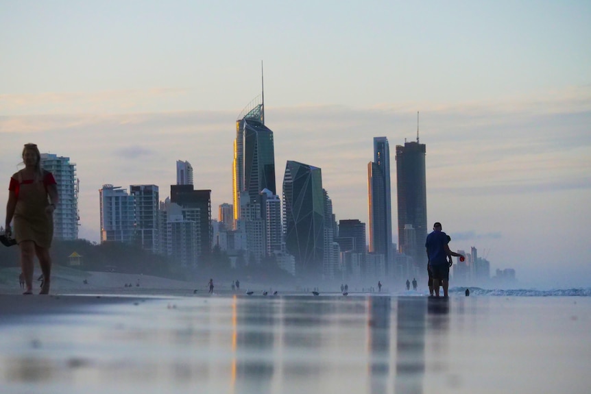 City skyline behind beach, two people walk in the foreground, many on the beach behind.