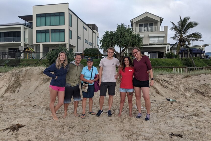 A group of six people stand on a stand dune, behind which stand large houses.