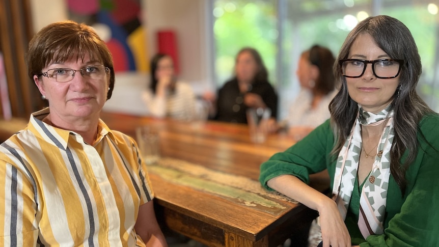 Mara and Litsa sit at the front of a table with serious expressions, while three women are unidentified and blurry behind them.