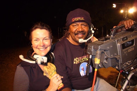 Elton Rosas and unknown woman wearing headphones looking to camera and standing next to studio camera.