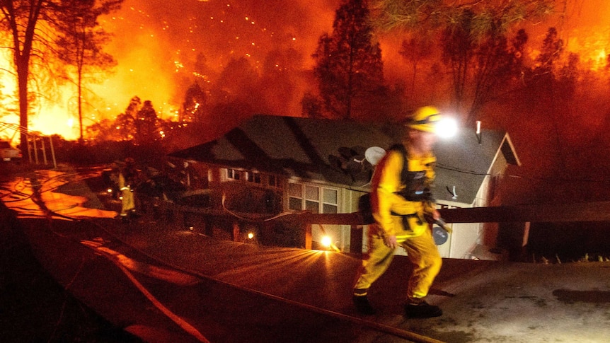 A firefighter walks up a hill in front of a house threatened by high flames which burn through trees