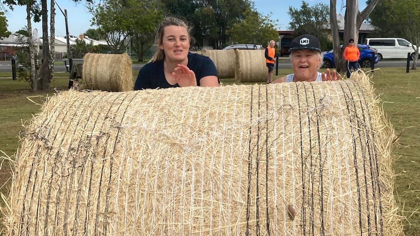 Two women laugh as they push a very large round hay bale toward the camera.