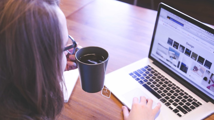 A woman drinks a cup of tea while working on a laptop.