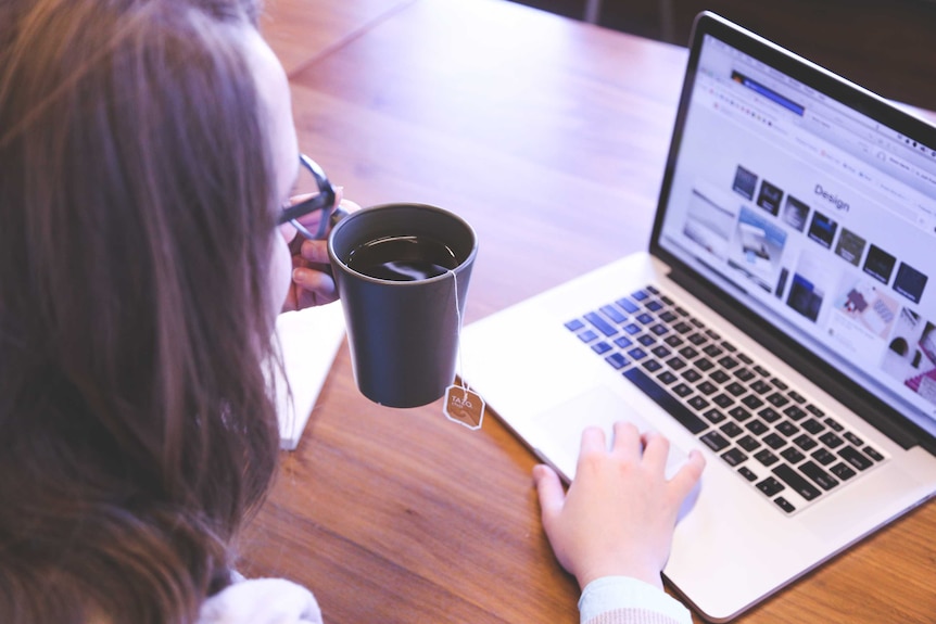 A woman drinks a cup of tea while working on a laptop.