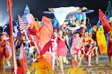 A woman lies on a paddleboard above the crowd as dancers re-enact a beach scene