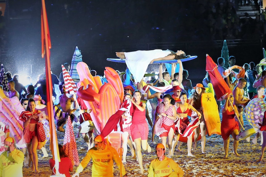 A woman lies on a paddleboard above the crowd as dancers enact a beach scene.