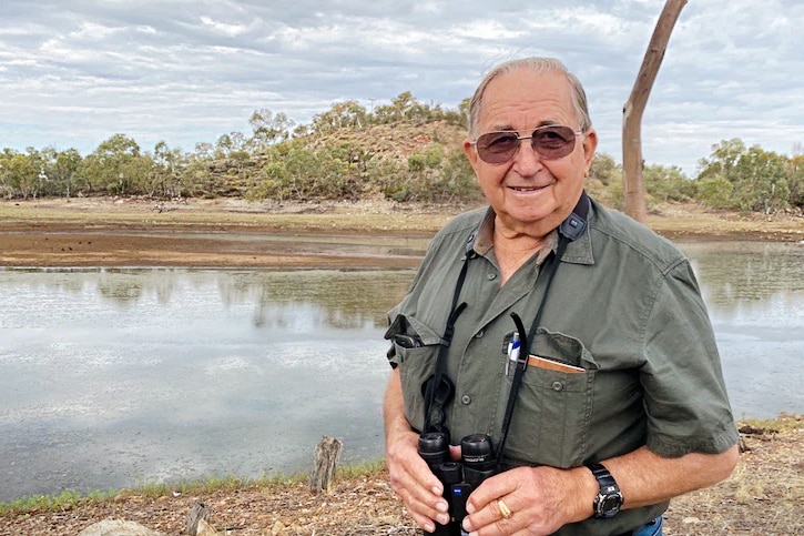 An older man stands in front of a lake holding binoculars