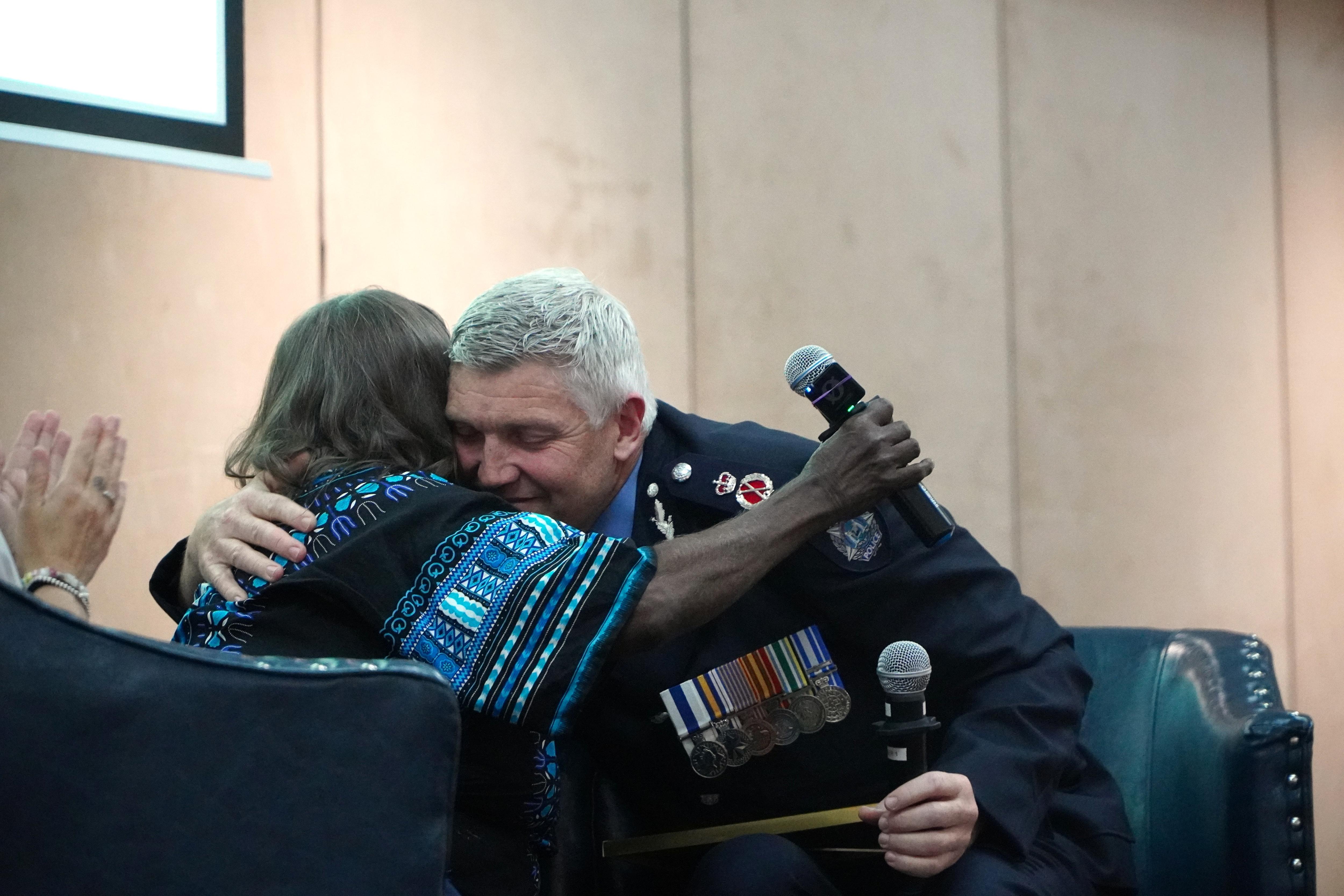 Col Blanch fica na frente da câmera enquanto abraça Daisy, que está segurando um microfone.