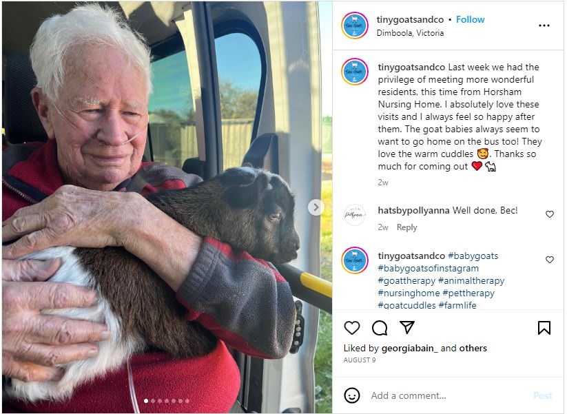 An older man wearing a thin tube across his face smiles as he holds a little brown and white goat.