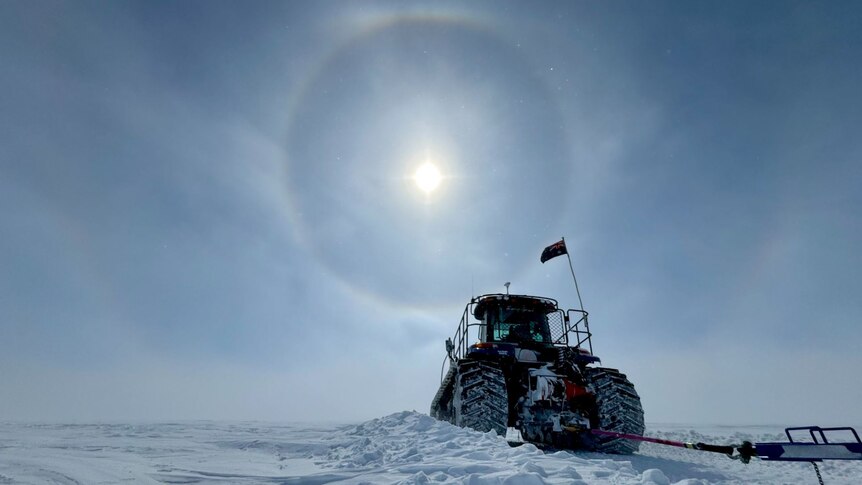 A black coloured tractor in the Antarctic snow with whispy clouds across the blue sky. A large halo surrounds the sun. 