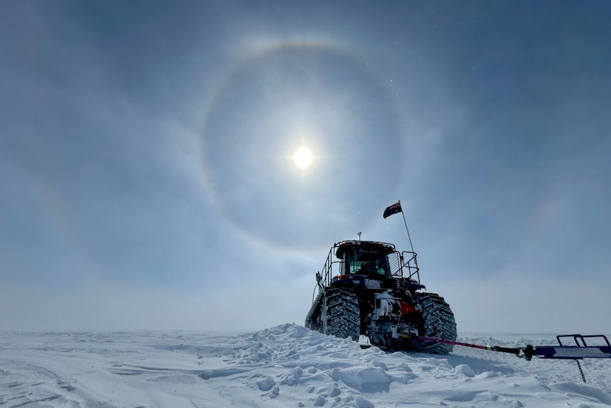 A black coloured tractor in the Antarctic snow with whispy clouds across the blue sky. A large halo surrounds the sun. 