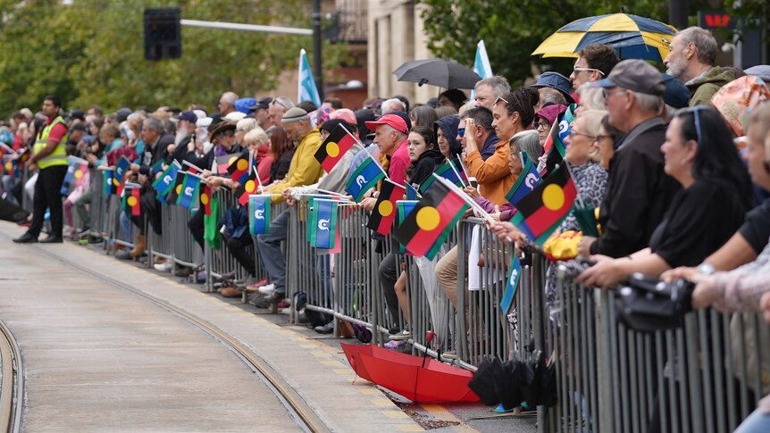 A crowd of people stand behind a gate, some waving Aboriginal and Torres Strait Islander flags